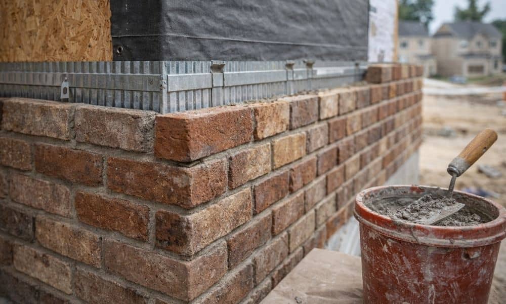 Close-up view of a masonry veneer brick wall under construction, showing brick texture, mortar bucket with trowel, and building frame details